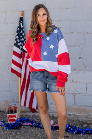Woman wearing a colorful sweatshirt with an American flag design, standing next to an American flag and Coca-Cola products.