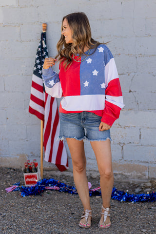 Woman wearing a colorful American flag shirt standing in front of a brick wall with an American flag.