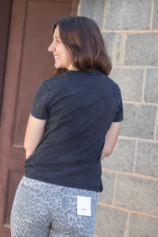 A woman shown from the back wearing a dark charcoal gray short-sleeve t-shirt and leopard-print jeans, standing against a gray brick wall and a brown wooden door.