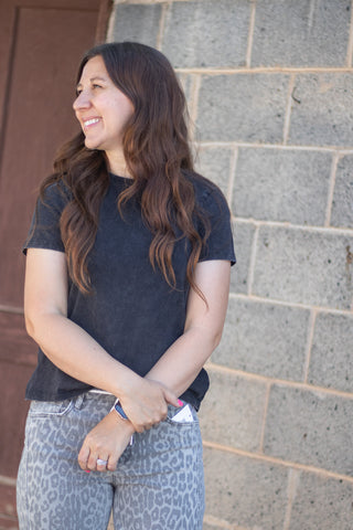 A woman with long brown hair stands in front of a gray brick wall, wearing a faded black t-shirt and gray leopard-print pants.