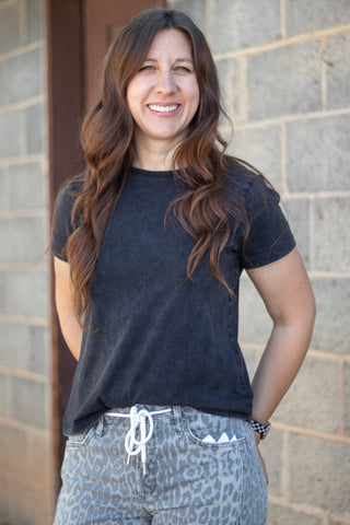 A smiling woman with long brown hair stands outdoors against a gray stone wall, wearing a faded black short-sleeve t-shirt and gray leopard-print pants with a white drawstring.