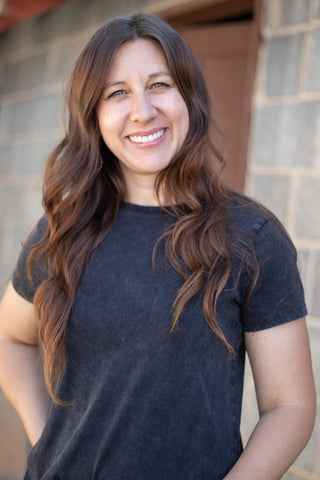 A smiling woman with long wavy brown hair wearing a dark charcoal gray t-shirt, standing in front of a gray brick wall.