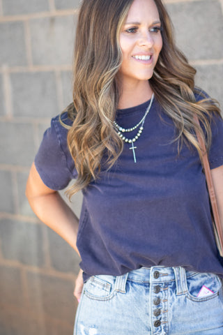 A woman wearing a dark navy blue t-shirt and light-wash denim jeans, accessorized with layered silver necklaces, standing against a gray brick wall.