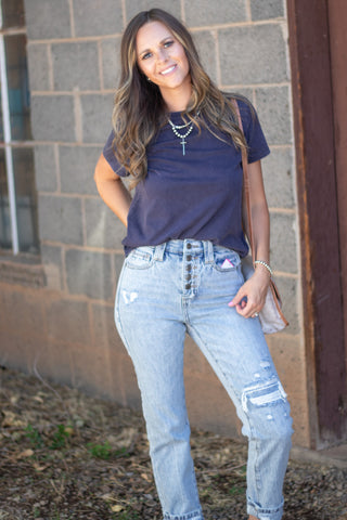 A woman posing outdoors in front of a brick wall, wearing a dark navy blue t-shirt, light-wash distressed denim jeans, layered silver necklaces, and a small beige shoulder bag.