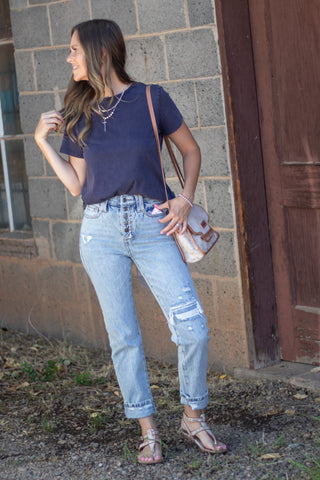 A woman wearing a navy blue t-shirt, light-wash distressed jeans, and metallic rose gold sandals, carrying a matching rose gold shoulder bag, standing outdoors against a brick wall and wooden door.