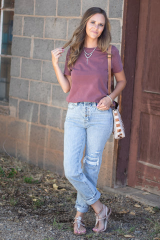 A woman stands outdoors in front of a stone wall and wooden door, wearing a muted mauve short-sleeve crewneck t-shirt, light-wash distressed jeans, and strappy flat sandals, with a woven crossbody bag.