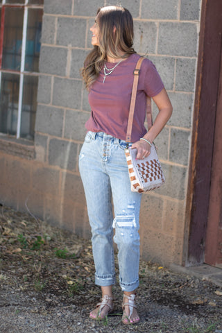 A woman stands outdoors against a gray brick wall wearing a mauve short-sleeve t-shirt, light-wash distressed jeans, and metallic sandals, while carrying a woven cream and brown crossbody bag.