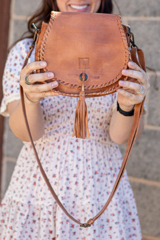 Brown leather satchel with tassel held by a person wearing a floral dress.