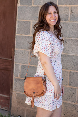 Brown leather satchel with tassel held by a person wearing a floral dress.