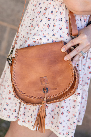Brown leather satchel with tassel held by a person wearing a floral dress.