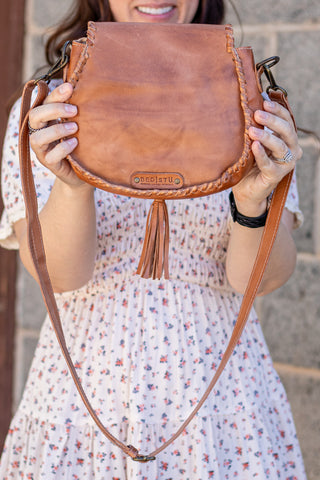 Brown leather satchel with tassel held by a person wearing a floral dress.