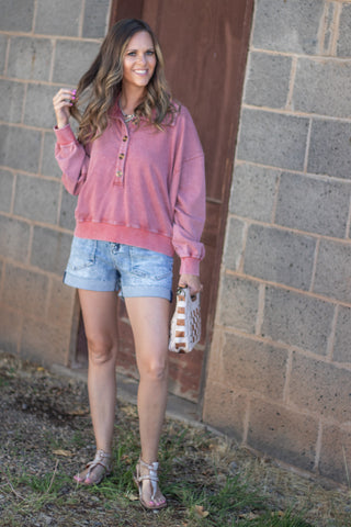 A woman stands outdoors in front of a gray cinderblock wall and brown door, wearing a dusty pink long-sleeve henley-style top, light blue denim shorts, and tan sandals, while holding a small woven clutch.
