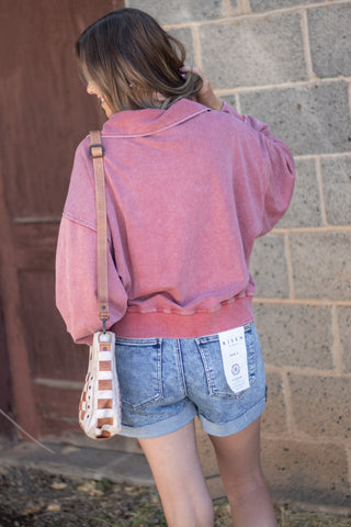 A woman shown from behind wearing a washed pink cropped sweatshirt and denim shorts, carrying a striped crossbody bag, standing against a gray brick wall.