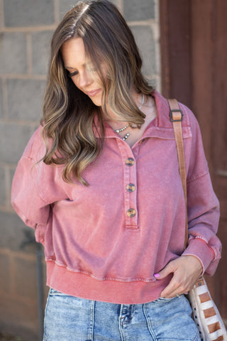 A woman wearing a dusty rose pink long-sleeve henley sweatshirt with a buttoned collar, paired with blue jeans and a striped shoulder bag, standing against a gray brick wall.