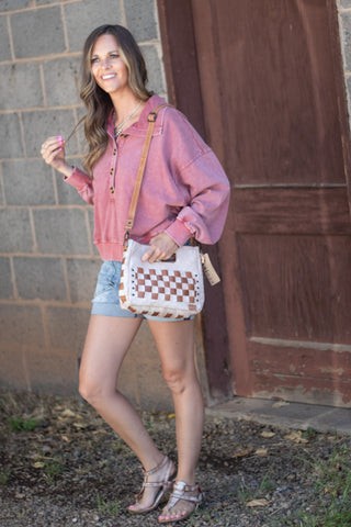 A woman wearing a pink zip-up hoodie, denim shorts, and metallic sandals is shown outdoors carrying a white woven crossbody bag with a brown checkerboard pattern and tan strap.