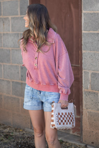 A woman wearing a pink washed henley sweatshirt and denim shorts, holding a white and brown checkered tote bag, standing in front of a gray brick wall.