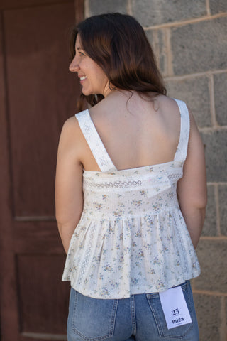 A woman shown from the back wearing a white sleeveless peplum top with a delicate blue and yellow floral print, paired with blue jeans. She is standing outdoors against a stone wall and a wooden door.