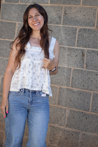 A woman with long brown hair smiling, wearing a white sleeveless floral top and blue jeans, standing against a gray stone wall.