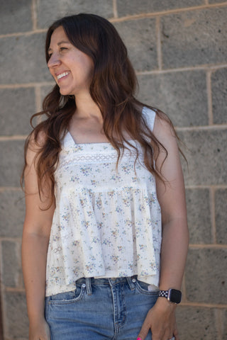 A woman with long brown hair smiling and looking to the side, wearing a sleeveless white top with a small blue and yellow floral print, paired with blue jeans, standing against a gray stone wall.