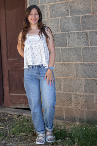 A woman stands outdoors in front of a gray brick wall and a brown wooden door, wearing a sleeveless white floral top, light blue jeans, and white slide sandals.