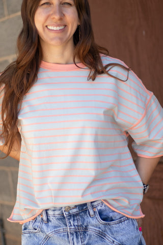 A woman wearing a light pink and white striped short-sleeve t-shirt with peach-colored trim, paired with blue jeans, standing against a brown wooden door background.