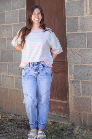A woman stands outdoors in front of a cinderblock wall and wooden door, wearing a white short-sleeve t-shirt with thin pink and light blue horizontal stripes, light-wash blue jeans, and white slide sandals.