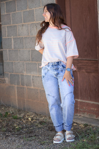 A woman wearing a light pink and white striped t-shirt with peach trim, light-wash blue jeans, and white slide sandals, standing outdoors against a gray brick wall and wooden door.