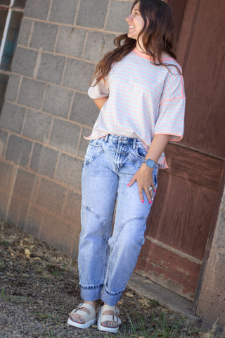 A woman wearing a light pink and white striped t-shirt, light-wash blue jeans, and white slide sandals, standing outdoors against a gray brick wall and a brown wooden door.