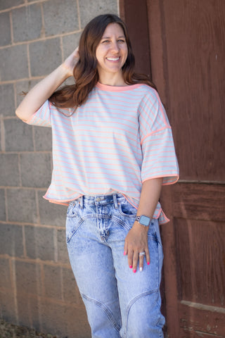 A woman wearing a light blue and pink striped short-sleeve t-shirt with light-wash blue jeans, standing outdoors next to a brown wooden door.