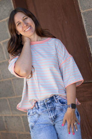 A woman wearing a light pink and white striped short-sleeve t-shirt with peach-colored trim, paired with blue jeans, posing in front of a rustic brown wooden door and stone wall.
