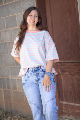 A woman wearing a white short-sleeve t-shirt with thin pink horizontal stripes and light blue jeans, standing in front of a gray brick wall and a brown wooden door.