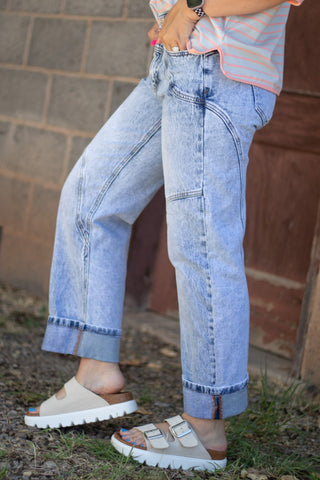 A person wearing light-wash blue denim jeans with a faded wash, paired with beige double-strap slide sandals and a pastel striped top, standing outdoors against a brick wall.