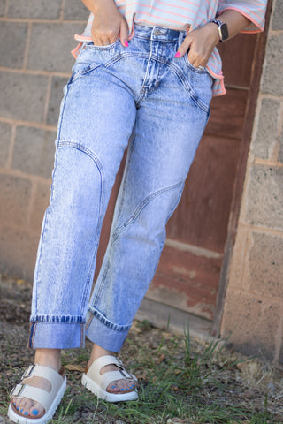 A person wearing light-wash blue denim jeans with contrast stitching and cuffed hems, paired with white slide sandals and a pink striped top, standing outdoors against a brick wall.