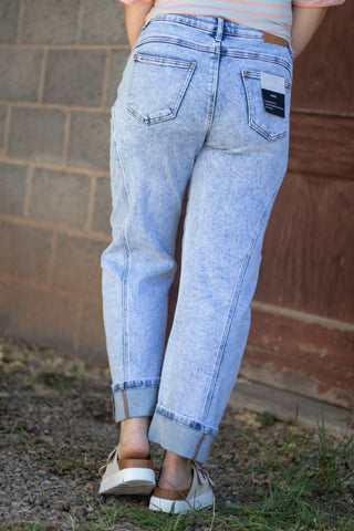 A person viewed from the back wearing light-wash blue cropped jeans with rolled cuffs, standing outdoors against a brick and wood wall.