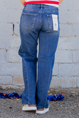 Person wearing blue jeans with a visible brand label, standing against a textured wall.