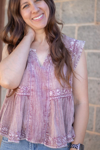A woman wearing a sleeveless dusty rose pink top with vertical textured stripes and embroidered details, paired with blue jeans, standing against a stone wall.