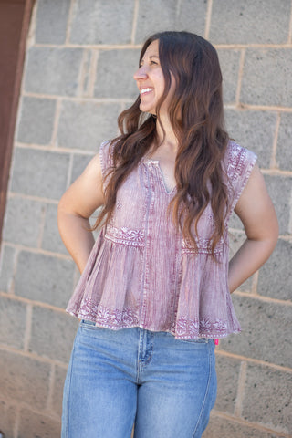 A woman wearing a sleeveless dusty purple peplum top with embroidered details and blue jeans, standing against a gray stone wall.