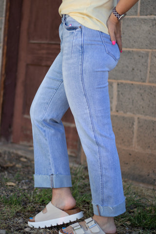 A person wearing light-wash blue cuffed jeans, a pale yellow top, and beige slide sandals, standing outdoors against a brick wall and wooden door.