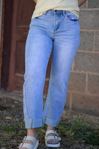 A person wearing light blue cropped jeans with rolled cuffs, paired with a pale yellow top and white slide sandals, standing outdoors against a brick wall and wooden door.