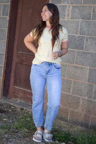 A woman wearing a pale yellow t-shirt with pink trim, light-wash blue jeans, and white slide sandals, standing outdoors against a gray brick wall and a brown wooden door.