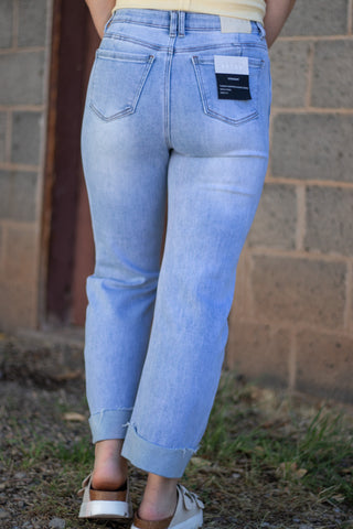 A person shown from the back wearing light-wash blue jeans with rolled cuffs, standing outdoors against a brick wall.