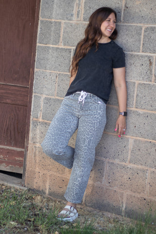 A woman wearing gray leopard-print sweatpants and a black t-shirt leans against a concrete block wall outdoors.
