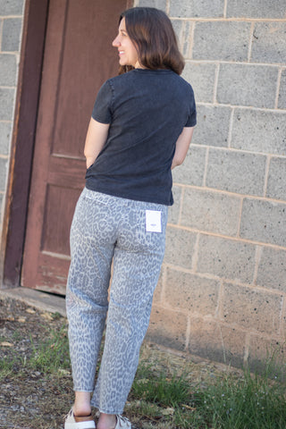 A woman wearing light blue-gray leopard print pants and a dark charcoal t-shirt, photographed from behind in an outdoor setting next to a cinderblock wall.