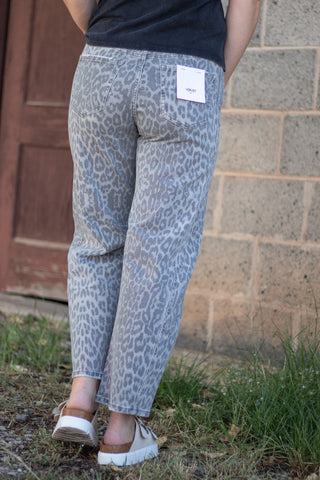 A person shown from the back wearing light gray leopard-print pants, a black top, and white sneakers, standing outdoors against a brick wall and wooden door.