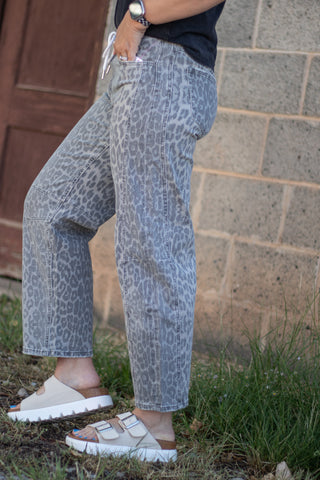 A person wearing gray leopard-print jeans, white slide sandals, and a black top, standing outdoors against a stone wall.