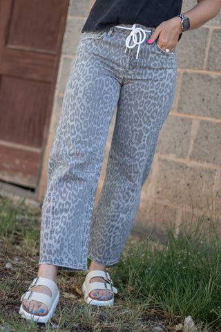 A person wearing light gray leopard-print jeans with a white drawstring waist, paired with white slide sandals, standing outdoors against a brick wall and wooden door.