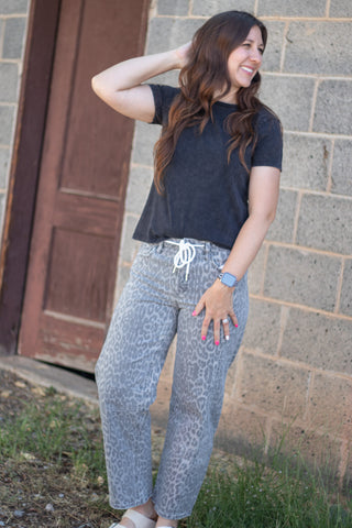 A woman wearing a black t-shirt and gray leopard-print sweatpants stands outdoors against a stone wall and wooden door.