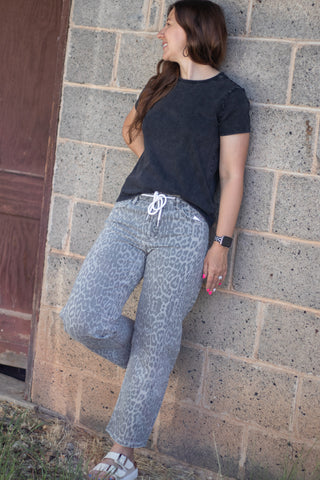 A woman wearing gray leopard-print pants and a black t-shirt leans against a concrete block wall outdoors.