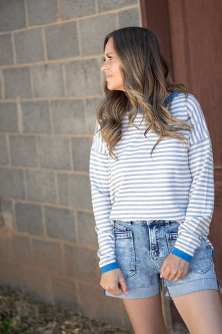 A woman wearing a blue and white striped long-sleeve crop top and denim shorts, standing outdoors against a gray brick wall.