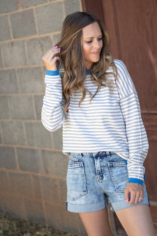 A woman wearing a blue and white striped long-sleeve shirt and denim shorts, standing outdoors against a gray brick wall and brown wooden door.